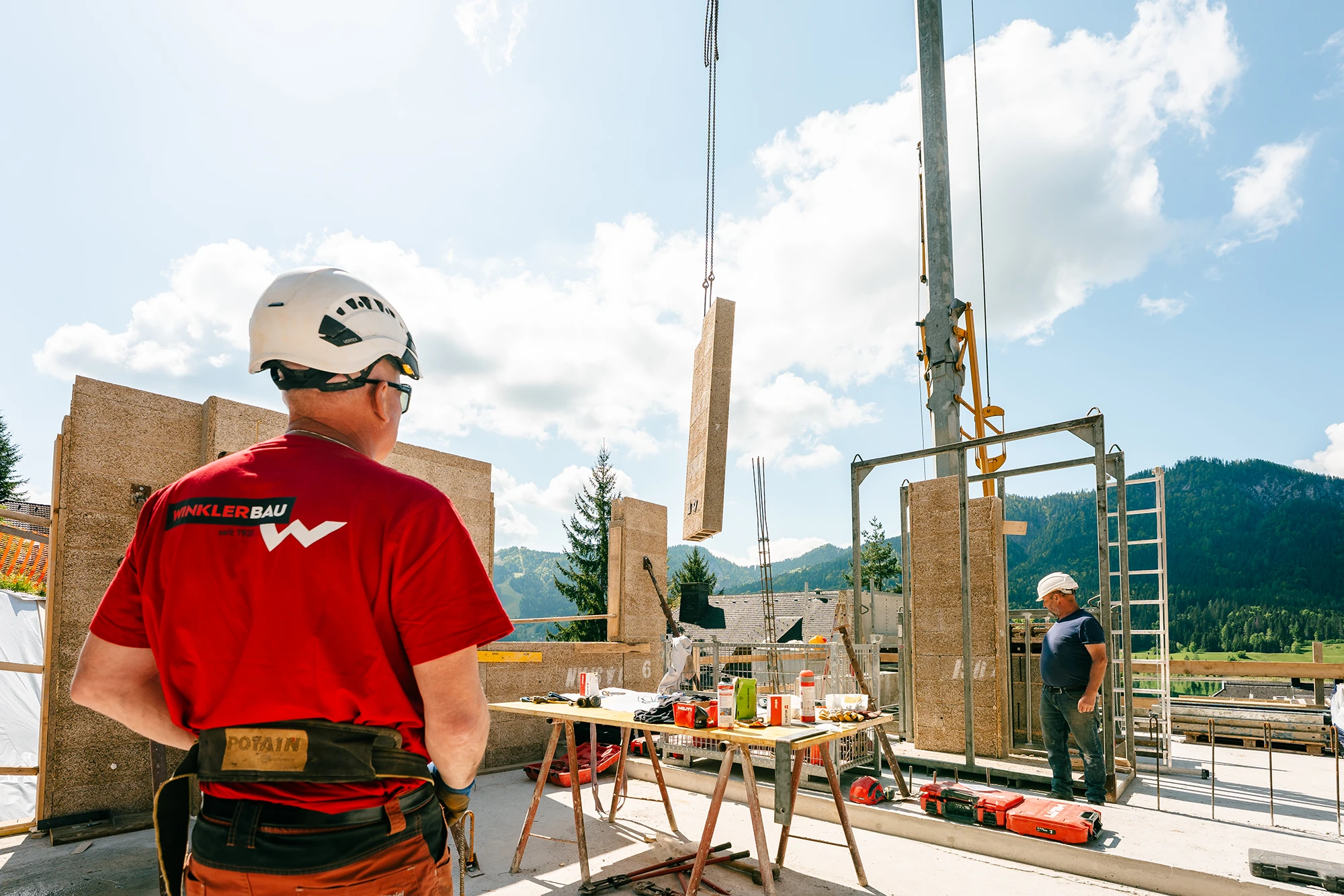 Zu sehen ist ein Mitarbeiter auf einer Baustelle der mit dem Kran ein Bauteil hochhebt.