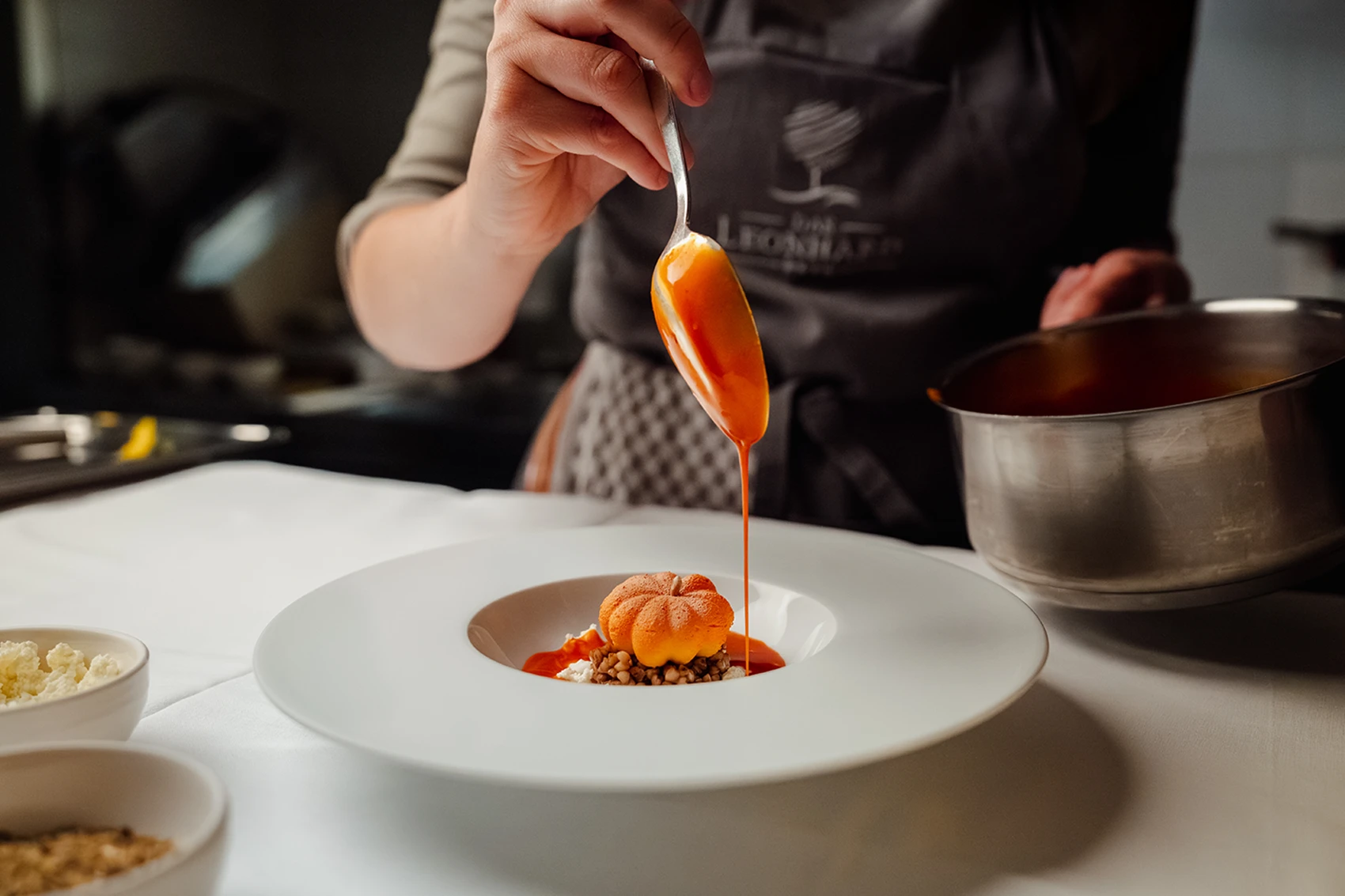 Jemand gießt Orangensauce über ein Gourmetgericht mit Getreide und einer kunstvoll geformten Garnierung und richtet das Essen an einem Tisch an, auf dem Schalen und ein Topf bereitstehen – vielleicht, um seinen automatisch gespeicherten Entwurf für eine elegante Tischpräsentation zu perfektionieren. – SquadTwenty Werbeagentur Kärnten