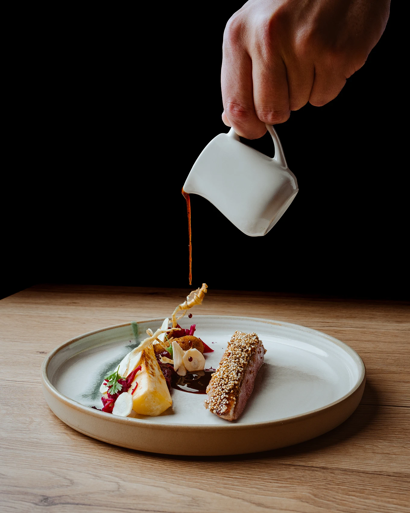 Auf dem Bild sieht man wie Sauce auf ein Teller mit Fisch und Gemüse eingegossen wird. Schwarzer Hintergrund und frische Farben.