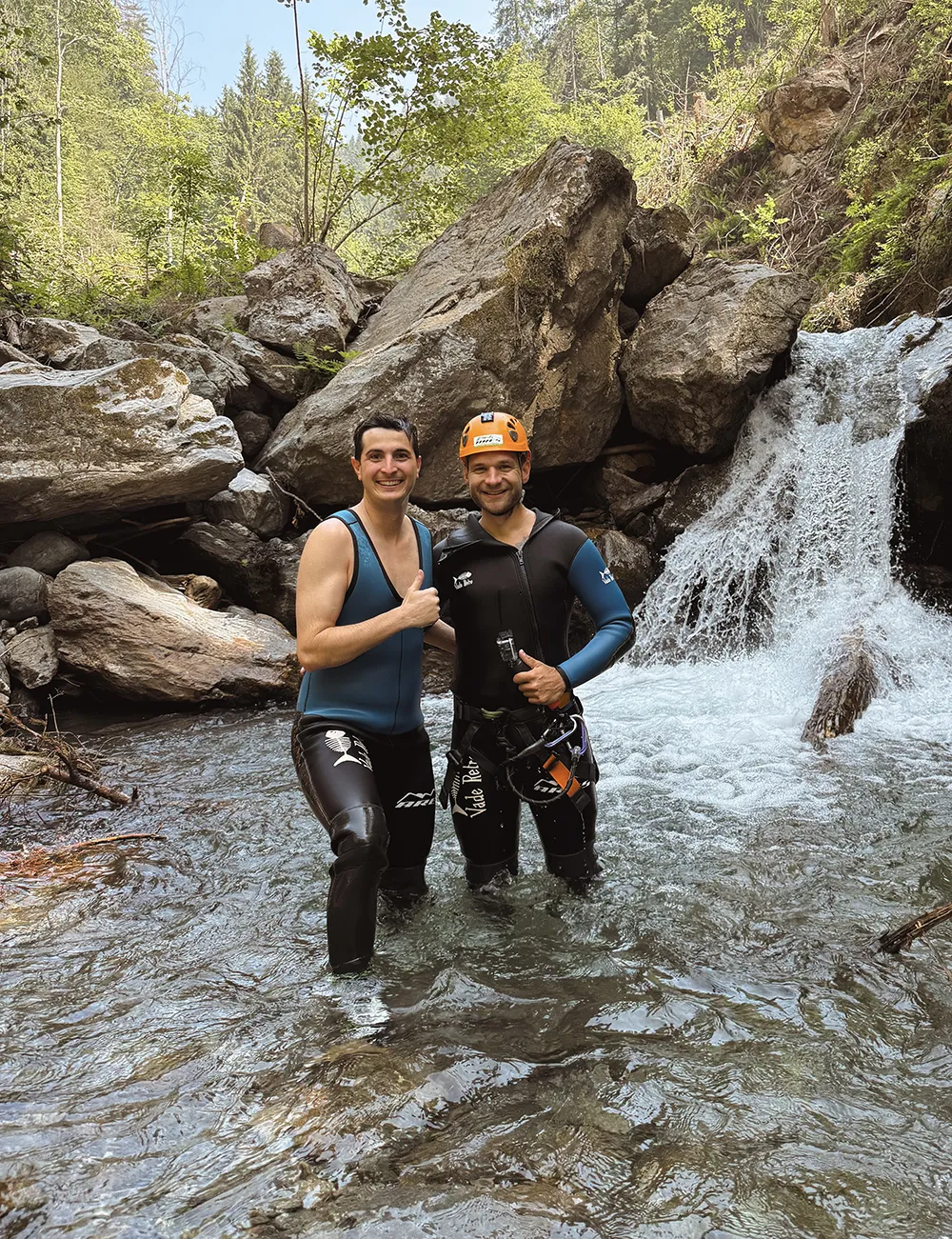 Valentino Zippo und Christofer Kemperle in Neoprenanzügen und Helmen stehen in einem felsigen Bach in der Nähe eines kleinen Wasserfalls, umgeben von Bäumen und Felsen. – SquadTwenty Werbeagentur Kärnten