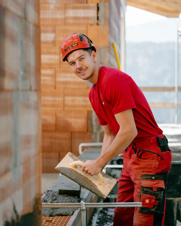 Es wird ein Maurer bei der Arbeiit gezeigt. Er steht vor der Wand und hält die Kelle in der Hand und lächelt in die Kamera.