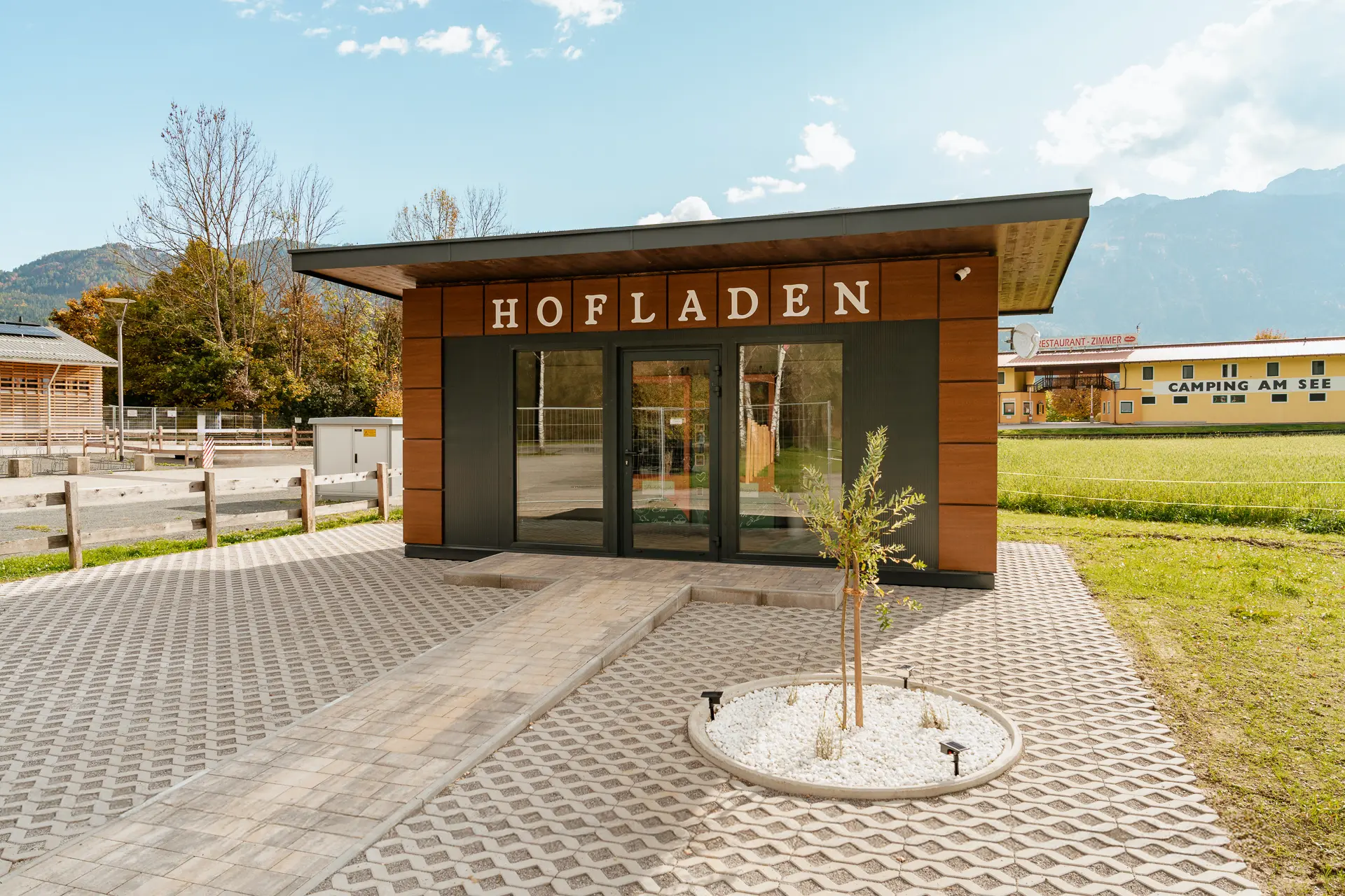 Ein kleines, modernes Gebäude mit der Aufschrift „Hofladen“ steht auf einer gepflasterten Fläche in der Nähe von Wiesen und Bergen unter einem teilweise bewölkten Himmel und lädt Besucher dazu ein, den Rasdorferhof zu erleben und den authentischen Charme eines Urlaubs auf dem Bauernhof zu entdecken.