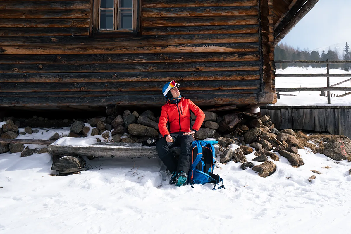 Eine Person in Winterkleidung sitzt auf einer Bank vor einer Holzhütte im Schnee und ruht sich aus, während ein blauer Rucksack neben ihr liegt – ein ruhiger Moment, wie ihn nur squadtwenty, eine auf Foto- und Videoproduktion in Kärnten spezialisierte Kreativagentur, einfangen kann. – SquadTwenty Werbeagentur Kärnten