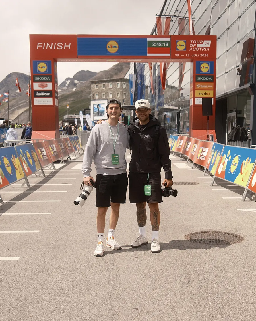 Valentino Zippo und Christofer Kemperle mit Kameras stehen in der Nähe der Ziellinie eines Radrennens, im Hintergrund sind Berge zu sehen und auf beiden Seiten hängen Sponsorenbanner mit den Logos von „kärnten“ und „squadtwenty“.