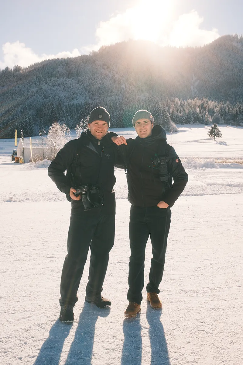 Zwei Personen in Winterkleidung stehen lächelnd mit Kameras auf verschneitem Boden vor einer sonnigen, schneebedeckten Berglandschaft – sie halten den Moment im Rahmen einer professionellen Foto- und Videoproduktion in Kärnten von squadtwenty fest. – SquadTwenty Werbeagentur Kärnten