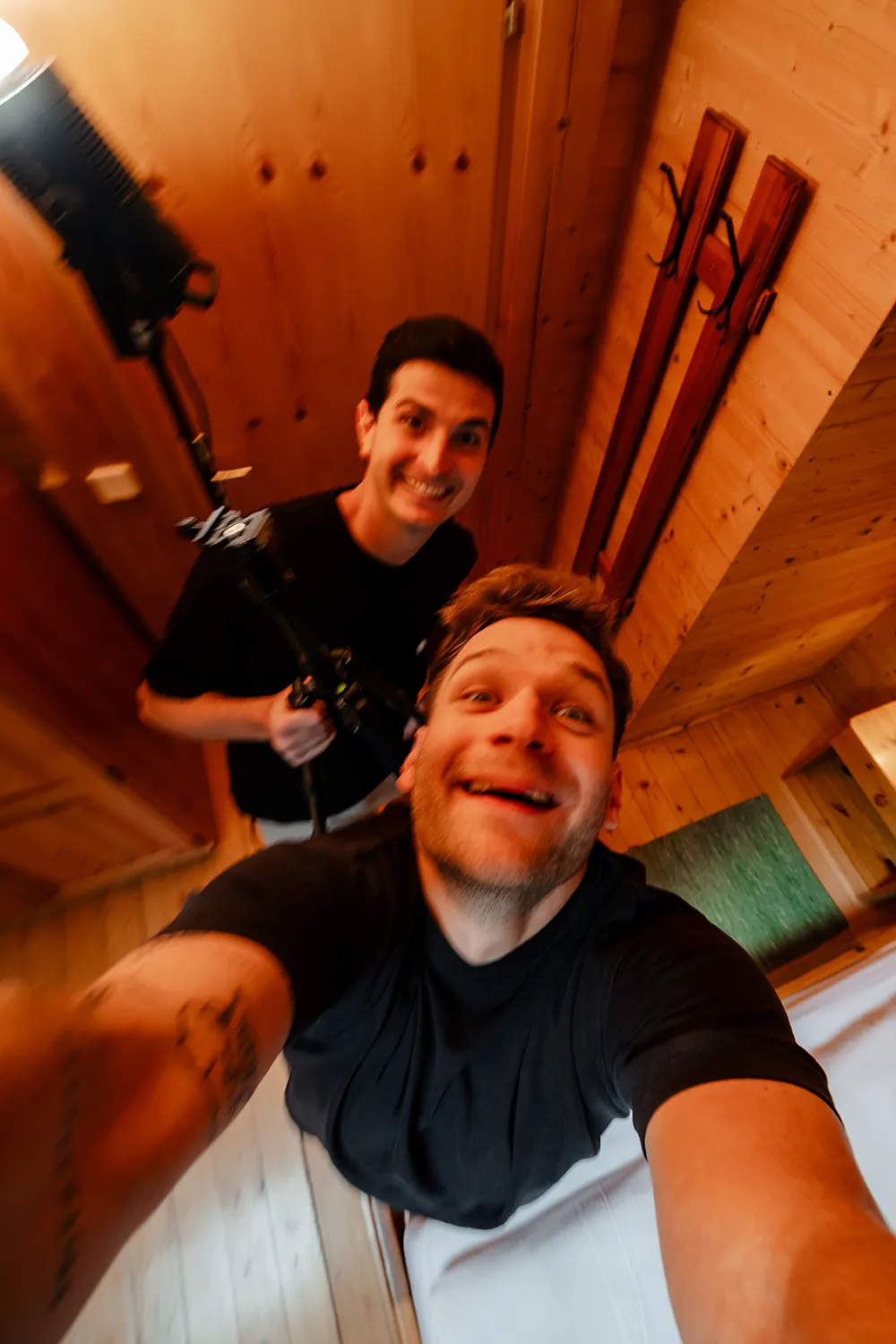 Zwei Männer lächeln in einem holzgetäfelten Raum; einer hält einen Beleuchtungsständer, während der andere aus der Vogelperspektive ein Selfie macht und so einen Blick hinter die Kulissen der neuesten Foto- und Videoproduktion ihres Teams in Kärnten festhält. – SquadTwenty Werbeagentur Kärnten