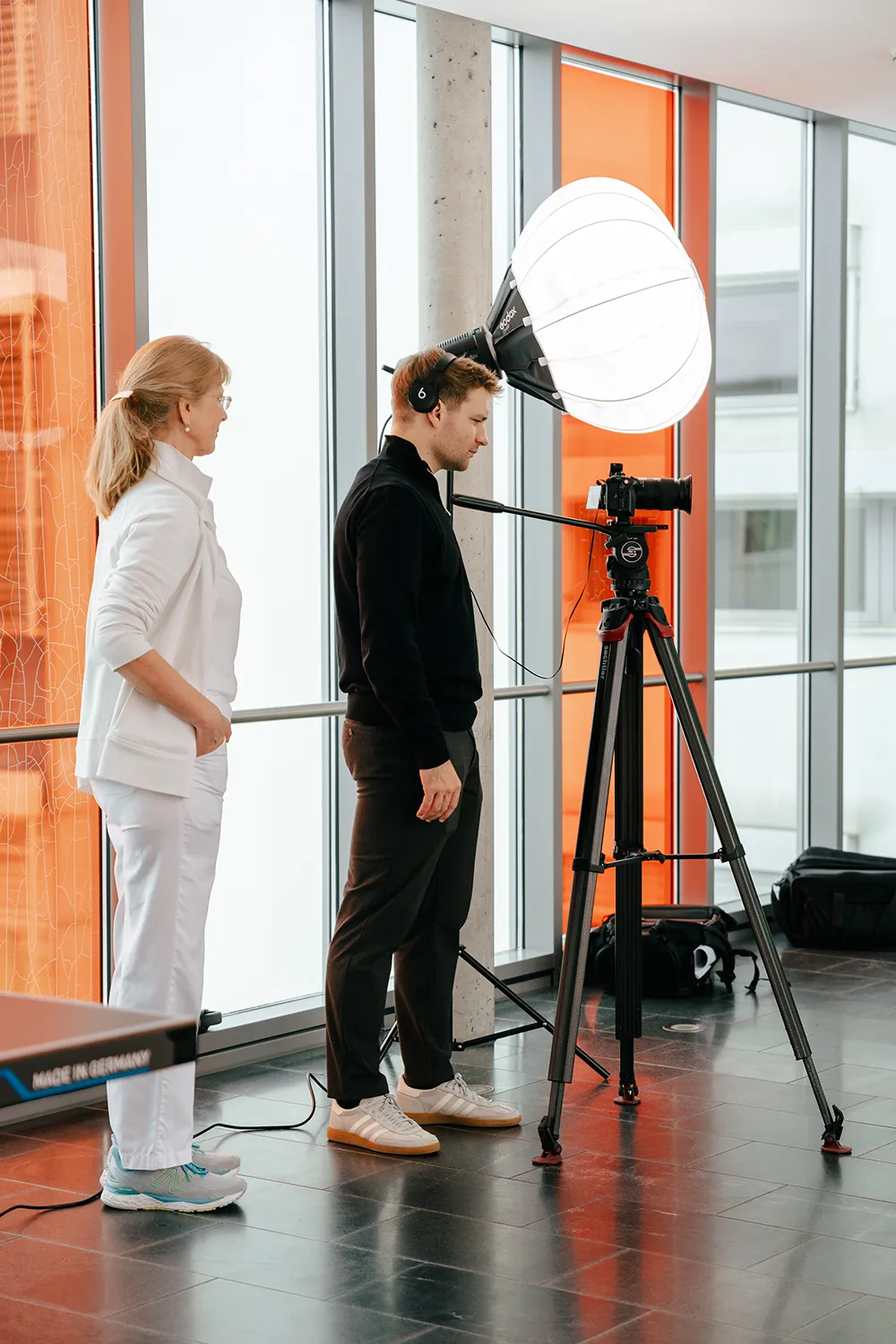 Zwei Personen stehen in einem Raum neben einer Kamera auf einem Stativ mit einem großen Ringlicht; eine davon, die „squadtwenty“ vertritt – eine Kreativagentur aus Kärnten, die sich auf Foto- und Videoproduktionen in Kärnten spezialisiert hat –, trägt Kopfhörer und bedient die Ausrüstung, während die andere aufmerksam zusieht. – SquadTwenty Werbeagentur Kärnten