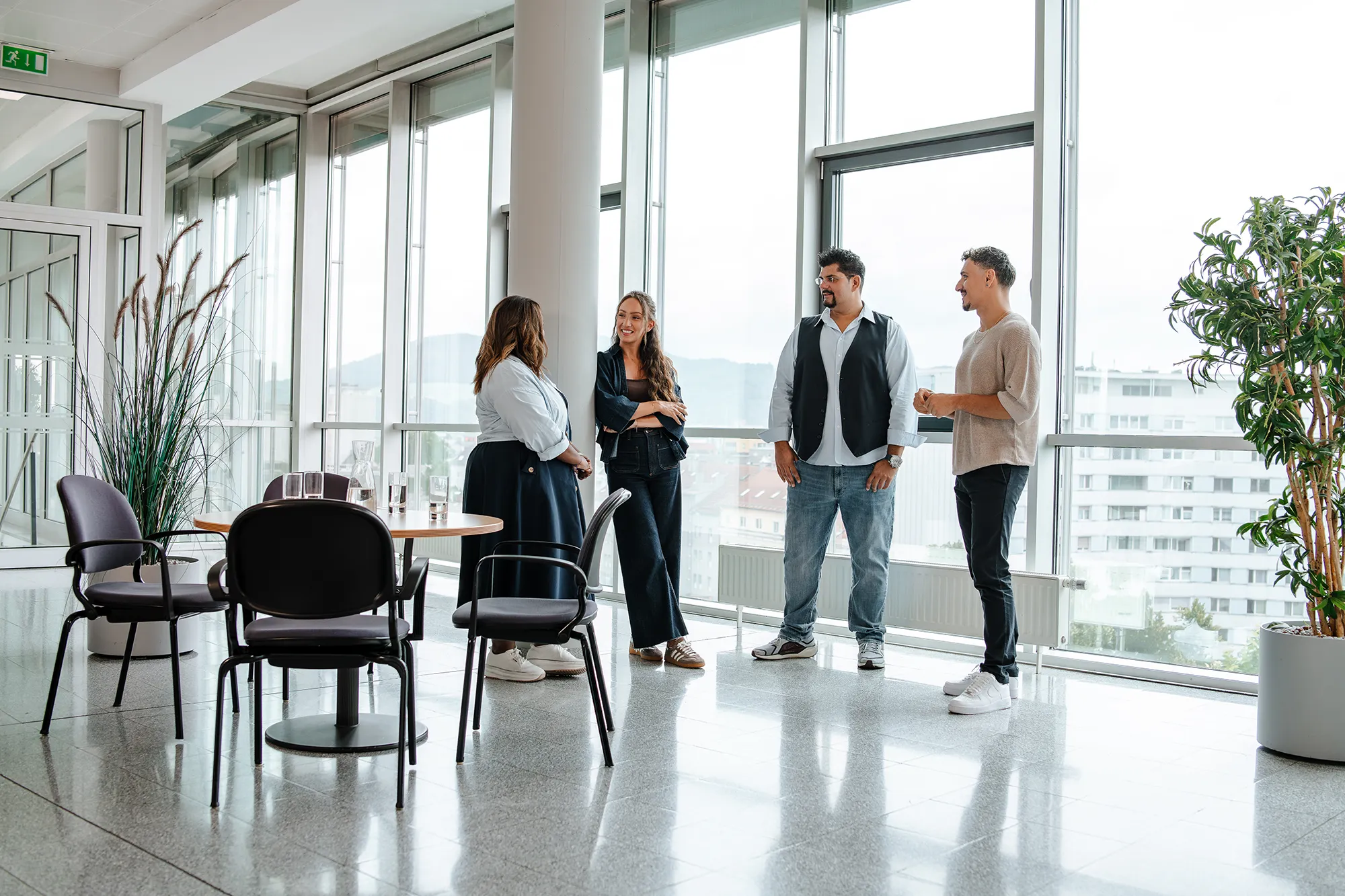 Ein vierköpfiges Team arbeitet an hellen Fenstern in einem eleganten Büro, an einem runden Tisch, mit digitalen Entwürfen auf Geräten in der Hand. – SquadTwenty Werbeagentur Kärnten
