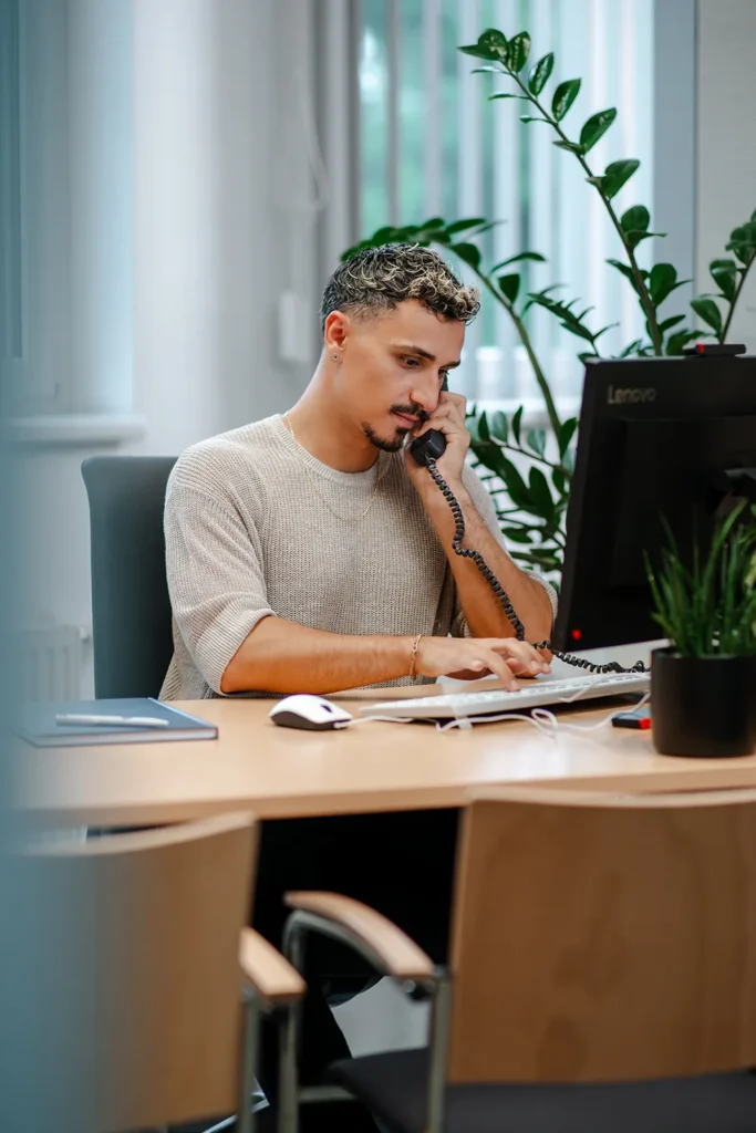 Ein Mann sitzt an einem Schreibtisch in einem Büro, spricht in ein schnurgebundenes Telefon und blickt auf einen Computermonitor, auf dem ein automatisch gespeicherter Entwurf zu sehen ist, umgeben von Pflanzen und Büromaterial. – SquadTwenty Werbeagentur Kärnten