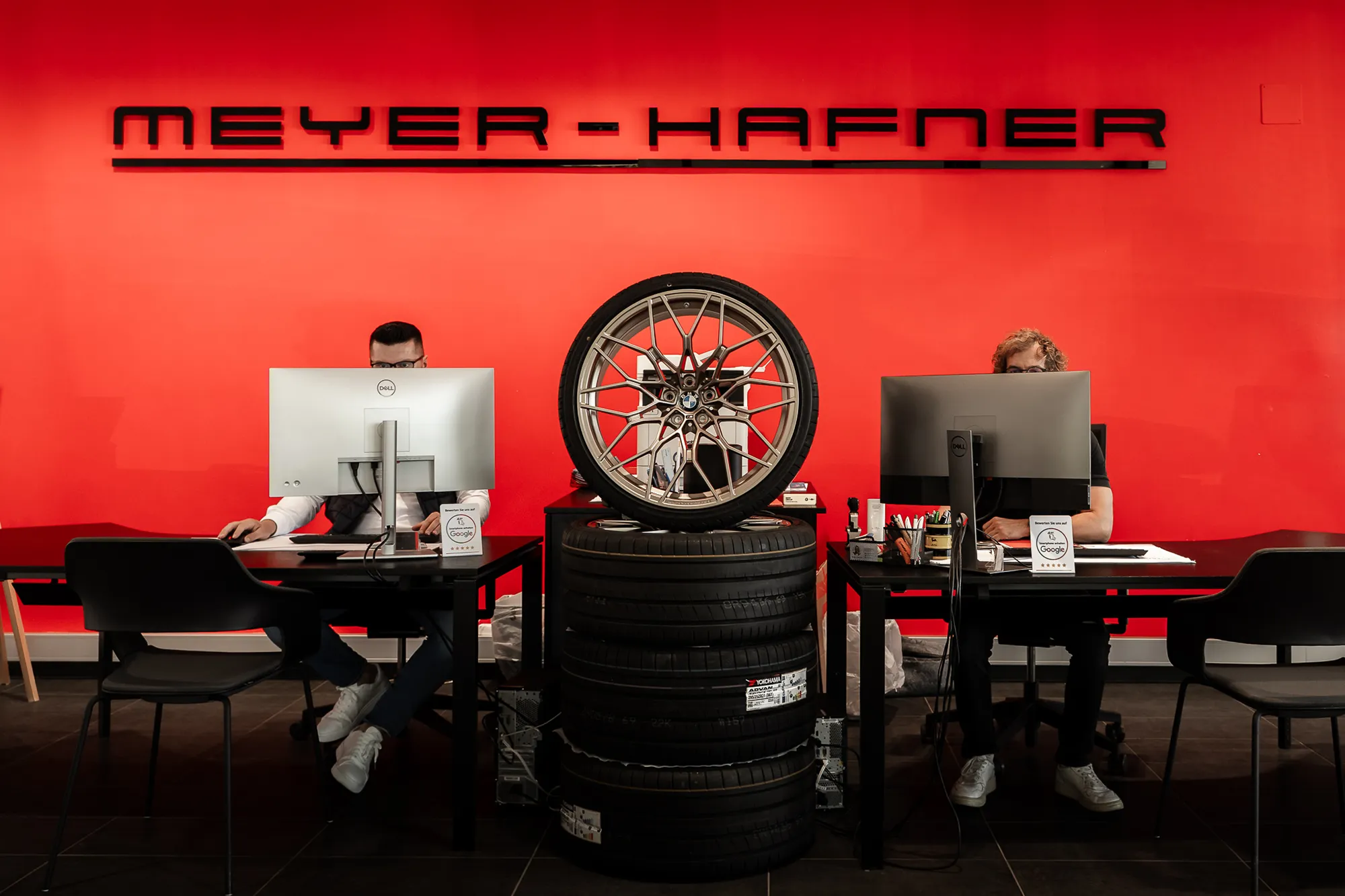 Zwei Personen arbeiten an Schreibtischen vor einer roten Wand mit der Aufschrift "MEYER-HAFNER". Zwischen ihnen steht ein Stapel Autoreifen mit einem Rad. – SquadTwenty Werbeagentur Kärnten