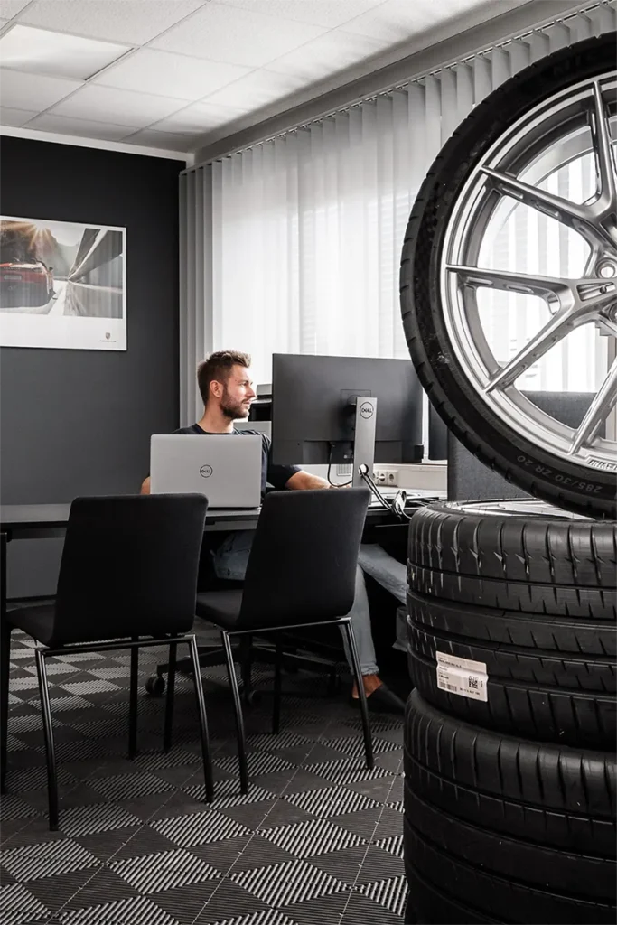 Ein Mann arbeitet an einem Schreibtisch mit einem Laptop und einem Monitor in einem modernen Büro. Im Vordergrund sind Stapel von Meyer-Hafner Autoreifen und ein großes Rad zu sehen. – SquadTwenty Werbeagentur Kärnten