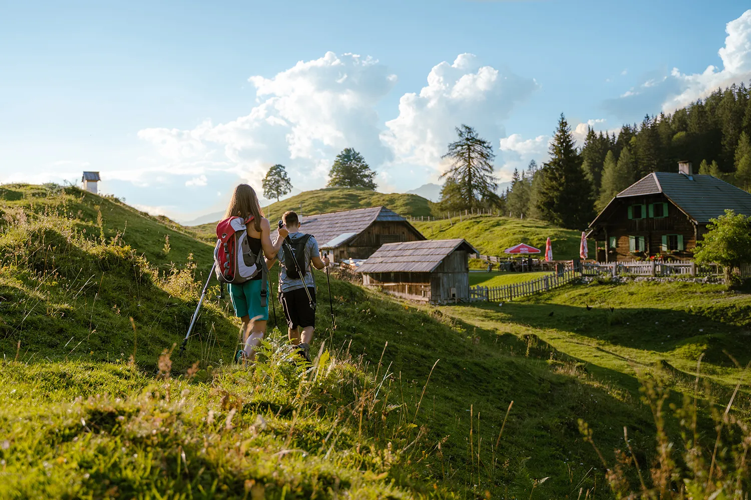 Abenteuerlustige Wanderer mit Rucksack wandern an einem üppigen Berghang in Richtung Berghütten - perfekt für auffällige Inhalte oder gewagte soziale Kampagnen. – SquadTwenty Werbeagentur Kärnten