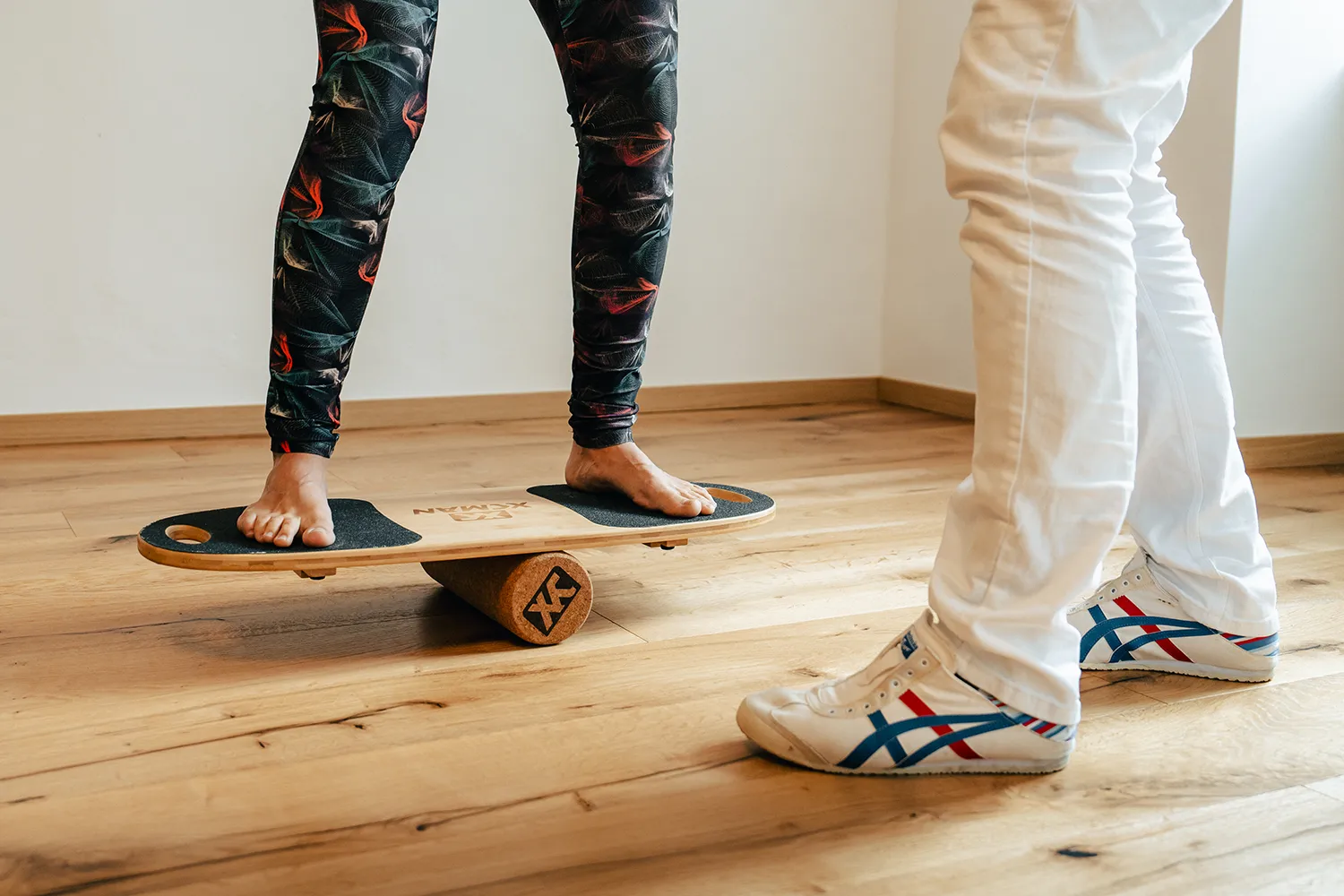 Ein barfuß laufender Mensch meistert ein hölzernes Balancierbrett, während ein Autor in weißer Hose und Turnschuhen auf dem glatten Holzfußboden zusieht. – SquadTwenty Werbeagentur Kärnten