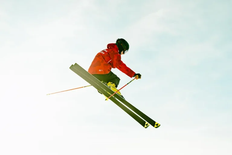 Skischule Weissensee: Ein Skifahrer in einer auffälligen roten Jacke startet mitten in der Luft, mit ausgefahrenen Skiern und Stöcken, vor einem minimalistischen Himmel. – SquadTwenty Werbeagentur Kärnten