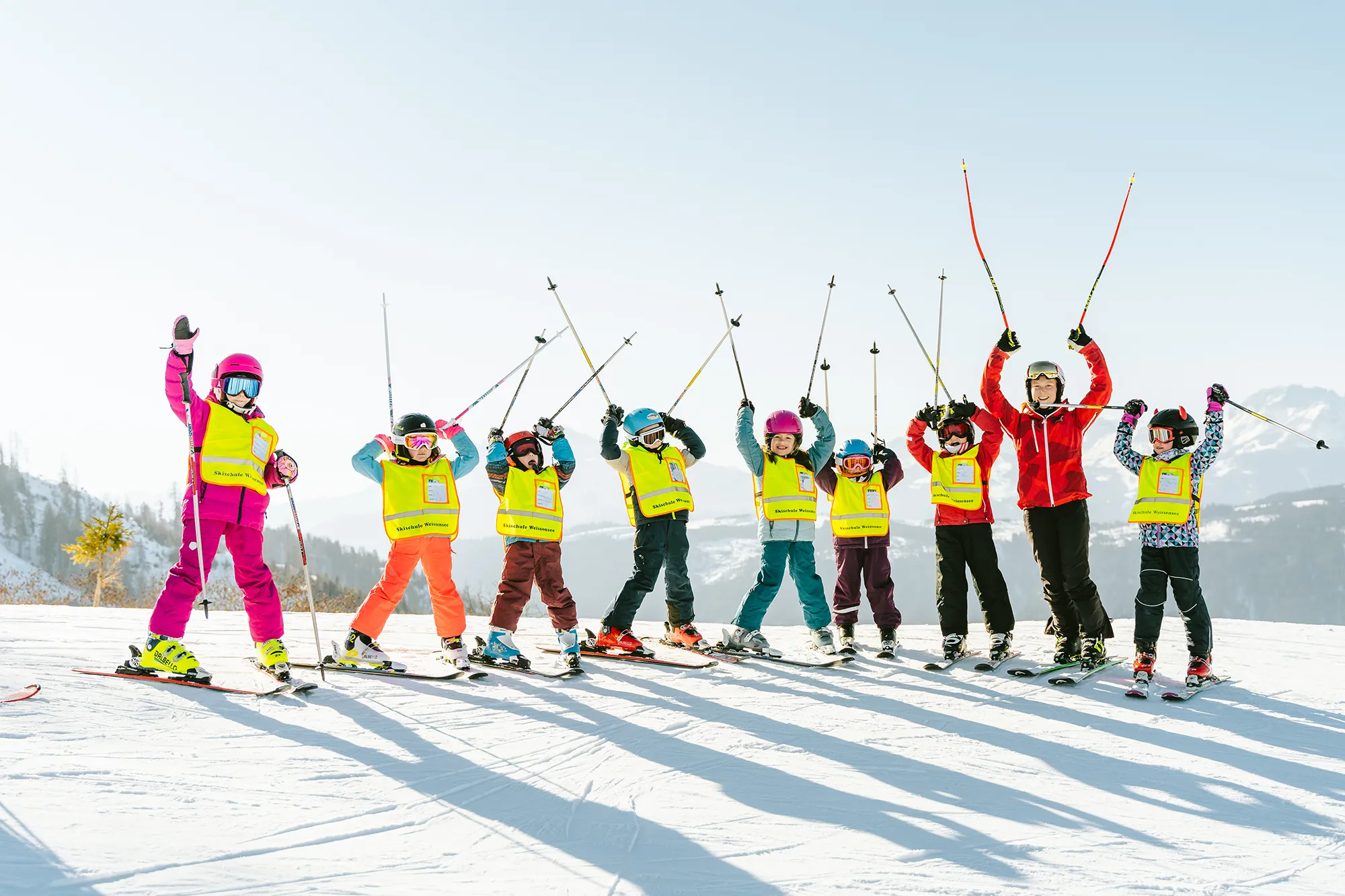 Kinder und ein Erwachsener in leuchtenden Winterklamotten und gelben Westen heben die Skistöcke auf einem verschneiten Hang in die Höhe - ideal für dynamische Markenvisualisierung. – SquadTwenty Werbeagentur Kärnten