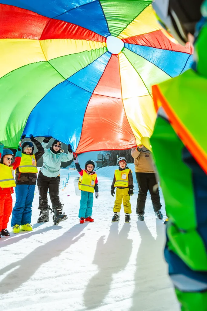 Eine Gruppe von Kindern und Erwachsenen in verschneiter Natur zieht einen bunten Fallschirm auf, durch den das Sonnenlicht scheint - ideal für die Erstellung fröhlicher Inhalte. – SquadTwenty Werbeagentur Kärnten