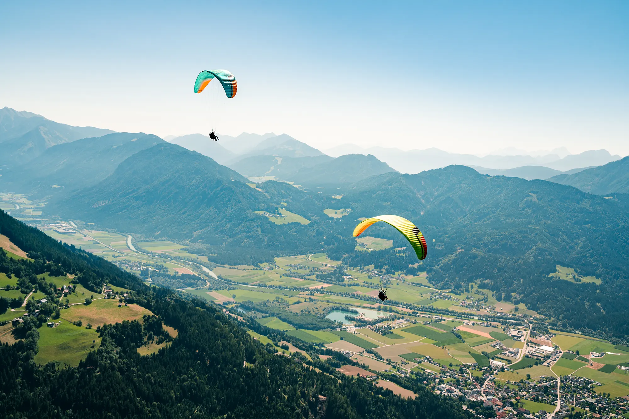 Heben Sie Ihre Marke hervor: Zwei Gleitschirme schweben über einem üppigen Tal und einem Bergpanorama und sorgen an einem strahlenden Sonnentag für soziales Aufsehen. – SquadTwenty Werbeagentur Kärnten