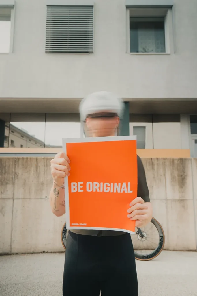 Eine Person mit verschwommenem Gesicht hält ein orangefarbenes "BE ORIGINAL"-Schild der Marke tubolito vor einem Gebäude und einem Fahrrad und fängt damit den radfahren Lifestyle ein. – SquadTwenty Werbeagentur Kärnten