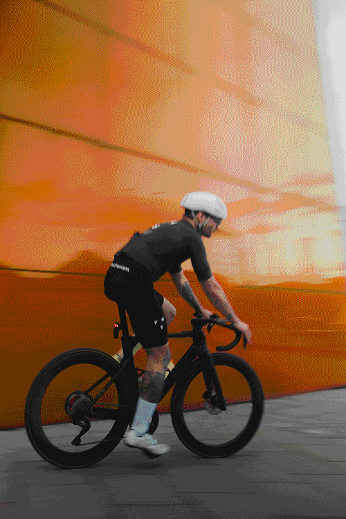 Ein Radfahrer mit Helm und Sportkleidung fährt auf einem schwarzen Fahrrad mit Tubolito-Schläuchen an einer orangefarbenen Wand vorbei. – SquadTwenty Werbeagentur Kärnten