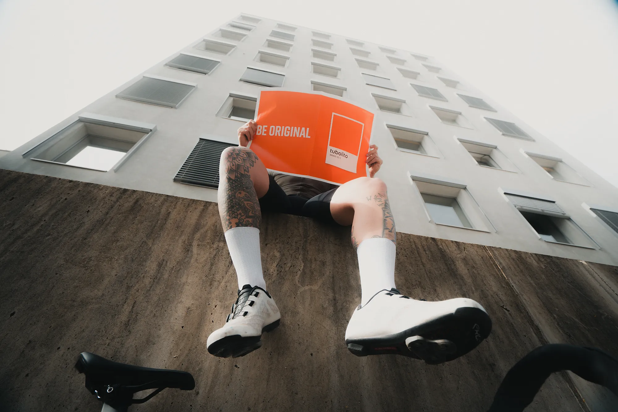 Eine Person posiert auf einem Felsvorsprung, die Beine schwingend, das Gesicht verdeckt durch ein fettes orangefarbenes "BE ORIGINAL"-Schild von tubolito, dahinter erhebt sich ein Stadtturm. – SquadTwenty Werbeagentur Kärnten