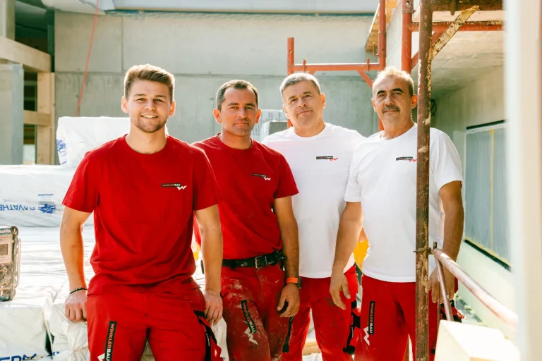 Vier Männer in rot-weißen Winklerbau Uniformen posieren selbstbewusst auf der Baustelle, aufgenommen für die Produktion neuer Inhalte an einem dynamischen Bauplatz. – SquadTwenty Werbeagentur Kärnten