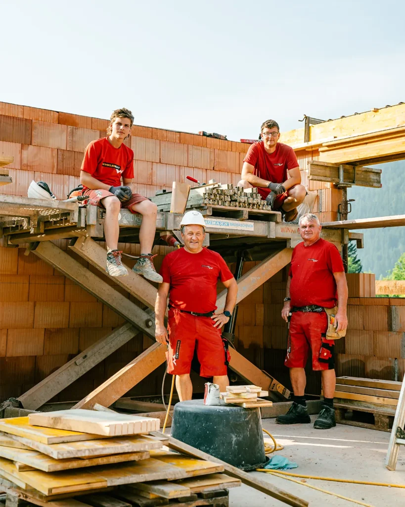 Vier Bauprofis in roter Montur stehen selbstbewusst auf der Baustelle - Ziegelwände, Gerüste und Werkzeuge zeigen das Know-how von Winkler Bau in Aktion. – SquadTwenty Werbeagentur Kärnten