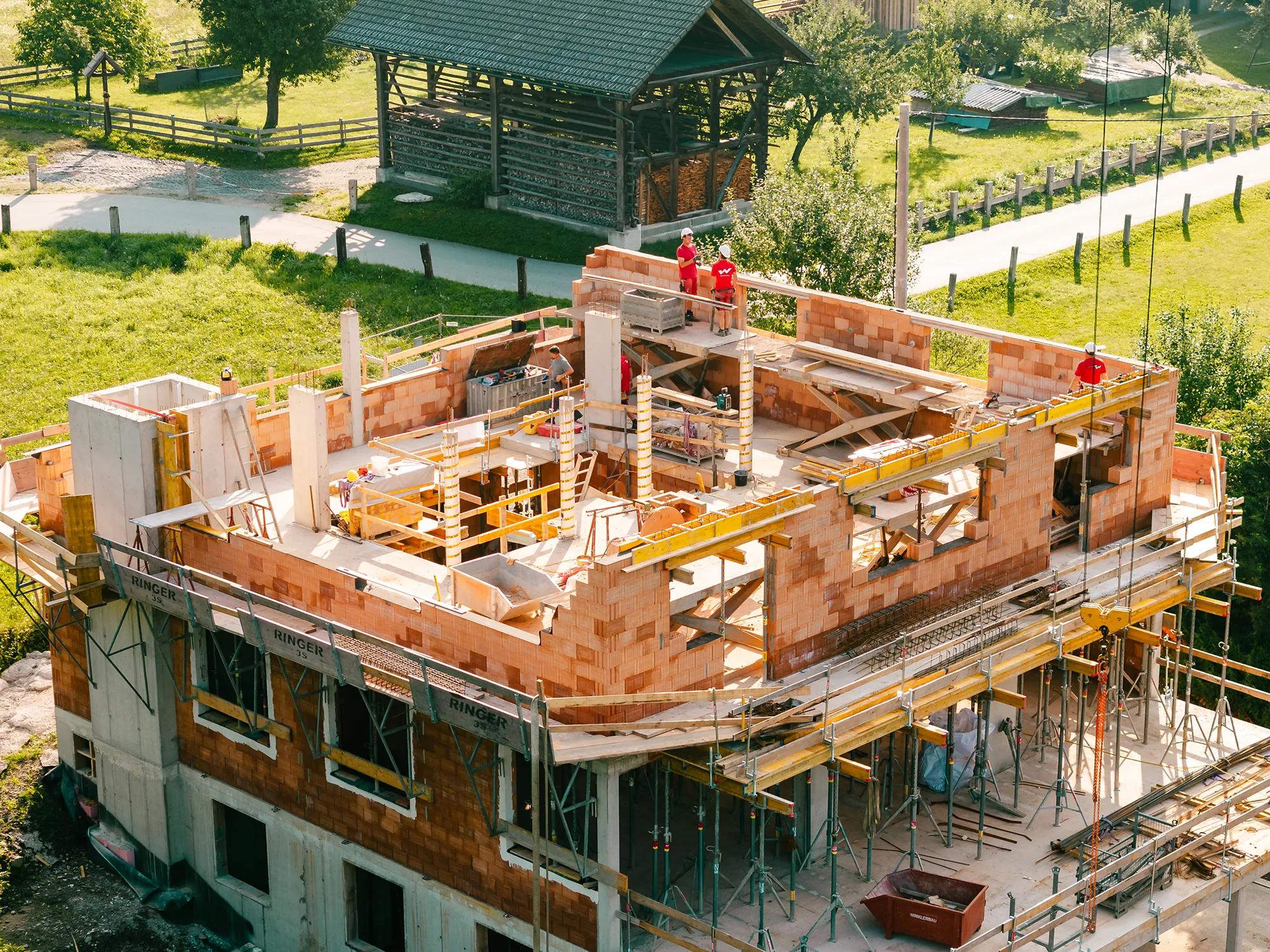Eine Winkler Bau-Baustelle mitten im Bau: Gerüste, Mauerwerk und behelmte Mitarbeiter vor grüner Kulisse - ideal für ein Corporate Shooting. – SquadTwenty Werbeagentur Kärnten