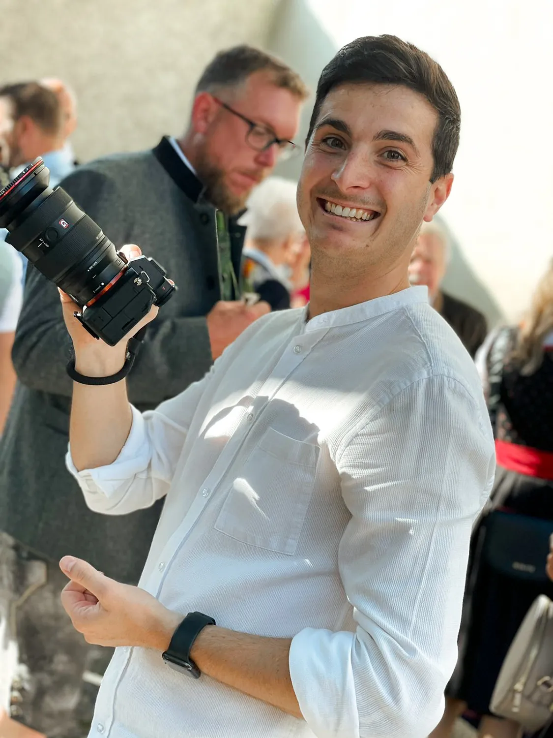 Valentino Zippo in einem weißen Hemd hält eine Kamera in der Hand und lächelt in die Kamera. – SquadTwenty Werbeagentur Kärnten