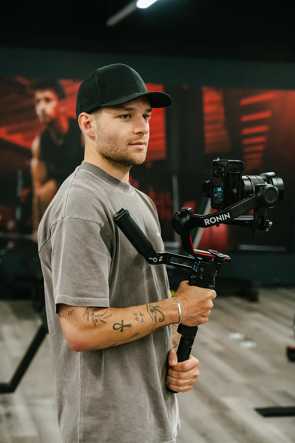 Ein Mann mit Mütze hält eine Kamera auf einem Ronin-Stabilisator in einem Fitnessstudio, mit unscharfen Fitnessgeräten und einem Plakat im Hintergrund - dynamische Inhalte für squadtwenty, Ihre kreative agentur kärnten für next-level branding. – SquadTwenty Werbeagentur Kärnten