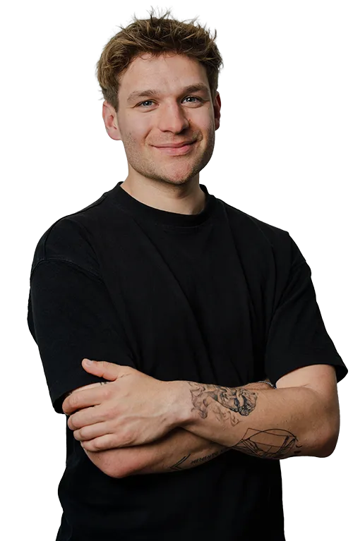 Person mit kurzen, hellbraunen Haaren und Tätowierungen auf dem linken Unterarm, Arme in einem schwarzen T-Shirt verschränkt, selbstbewusst lächelnd - SquadTwenty-Energie pur. – SquadTwenty Werbeagentur Kärnten