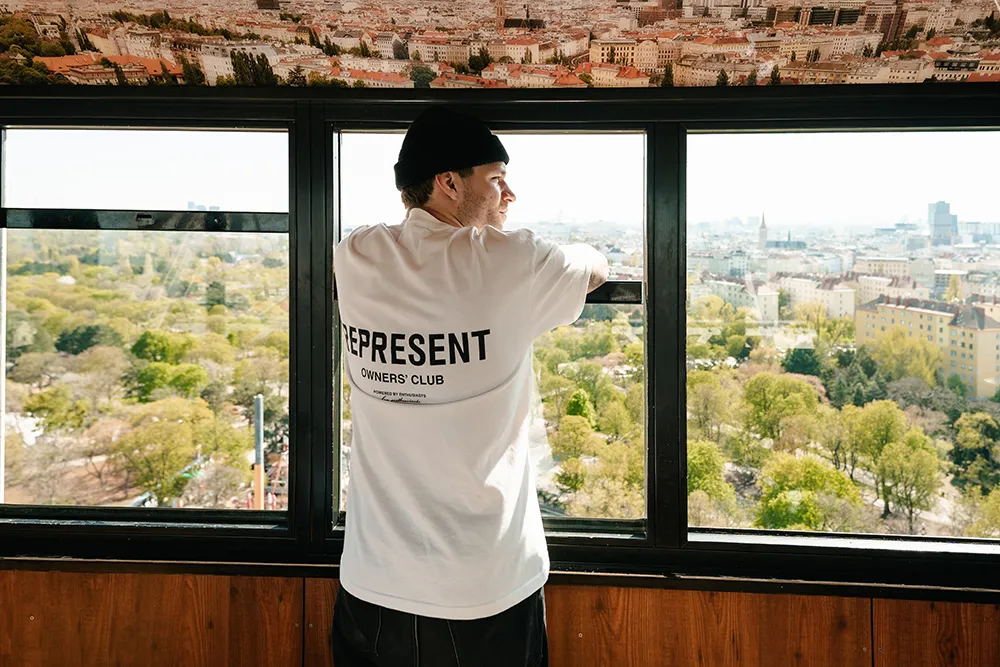 Eine Person mit einem "REPRESENT OWNER'S CLUB"-T-Shirt steht drinnen an einem großen Fenster und blickt auf eine Stadtlandschaft - eine inspirierende Szene für jede kreative agentur kärnten, die sich auf einzigartige Brandingprojekte konzentriert. – SquadTwenty Werbeagentur Kärnten