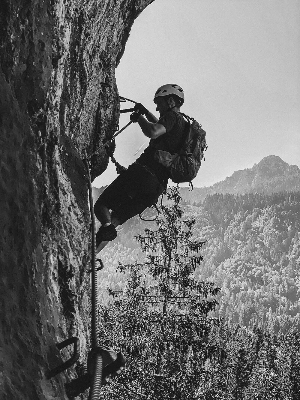 Eine Person, die einen Helm und einen Rucksack trägt, klettert mit Hilfe von Seilen und Sicherheitsausrüstungen auf eine felsige Klippe, mit bewaldeten Bergen im Hintergrund - perfekt für eine kreative agentur kärnten, die nach abenteuerlichen Bildern sucht. – SquadTwenty Werbeagentur Kärnten