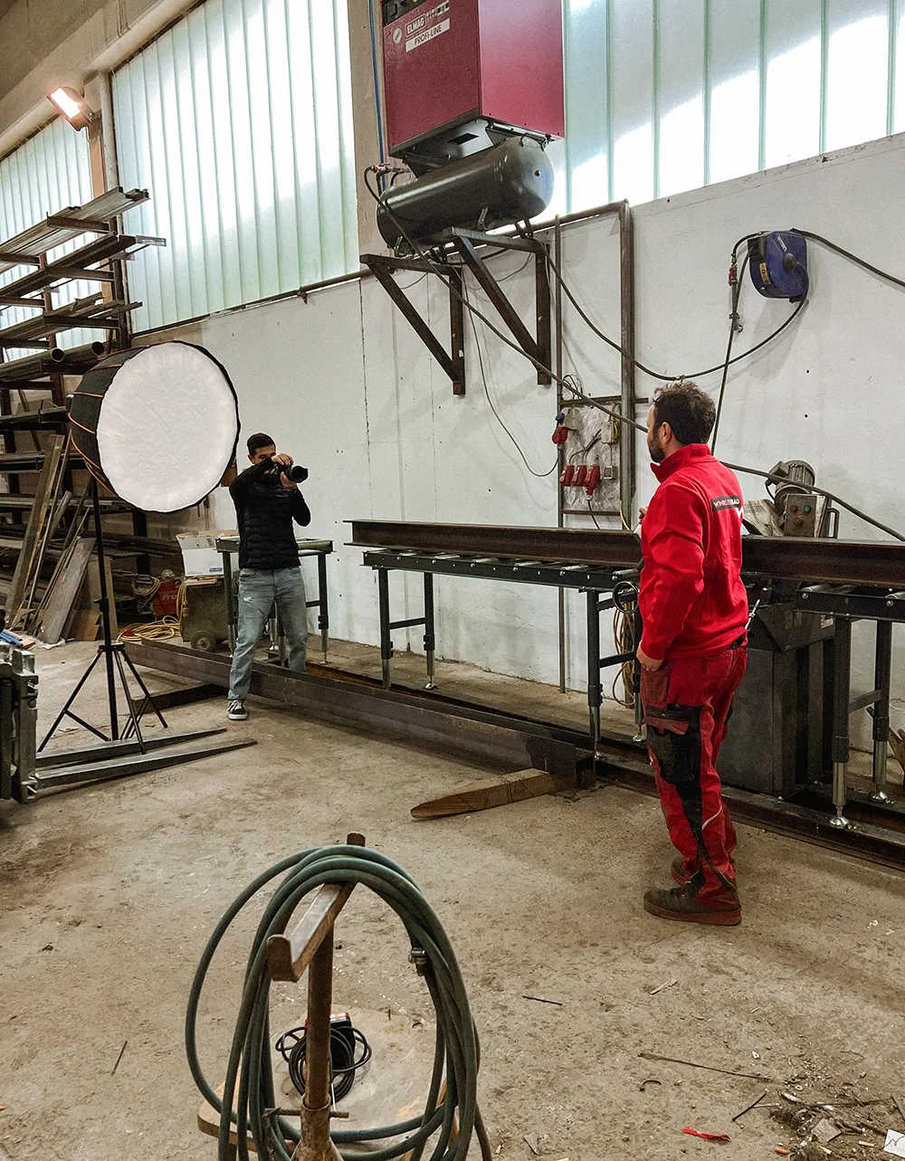 Ein Fotograf der Kreativagentur Kärnten fotografiert einen Mann in roter Arbeitskleidung in einer Industriehalle und setzt dabei professionelles Equipment und Licht für authentisches Branding Kärnten in Szene. – SquadTwenty Werbeagentur Kärnten