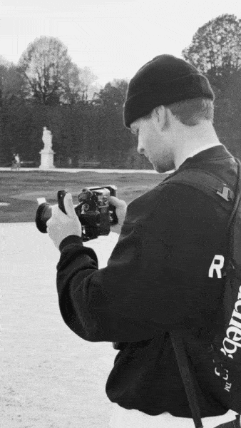 Eine Person mit Mütze und Rucksack steht im Freien und schaut nach unten, während sie eine Kamera in der Hand hält. Im Hintergrund sind Bäume und eine Statue zu sehen, die den kreativen Geist eines social media agentur kontakt-Moments einfangen. – SquadTwenty Werbeagentur Kärnten